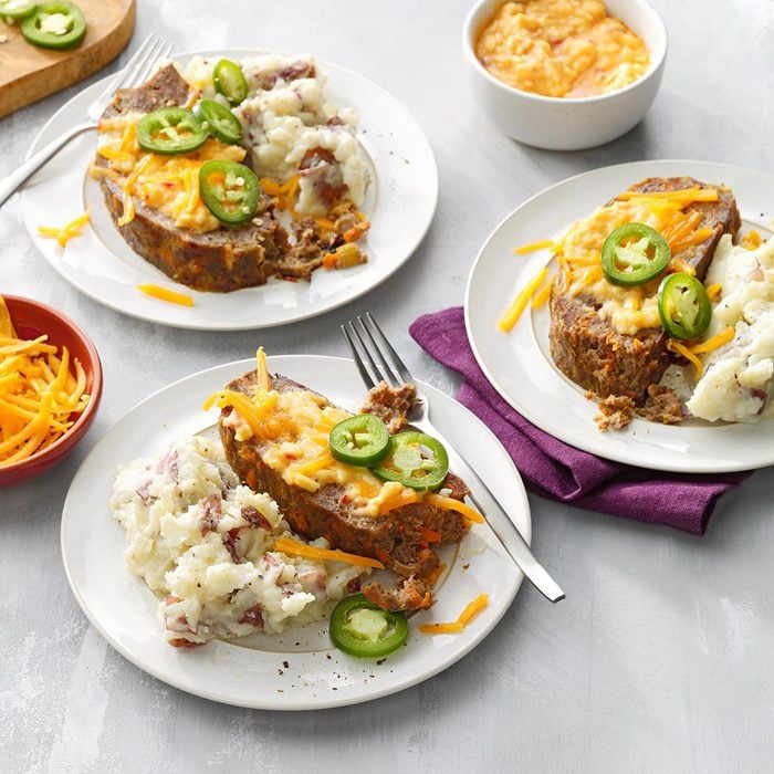 Meatloaf topped with cheese and jalapeÃ±os, paired with mashed potatoes, served on white plates with forks, set on a grey surface.