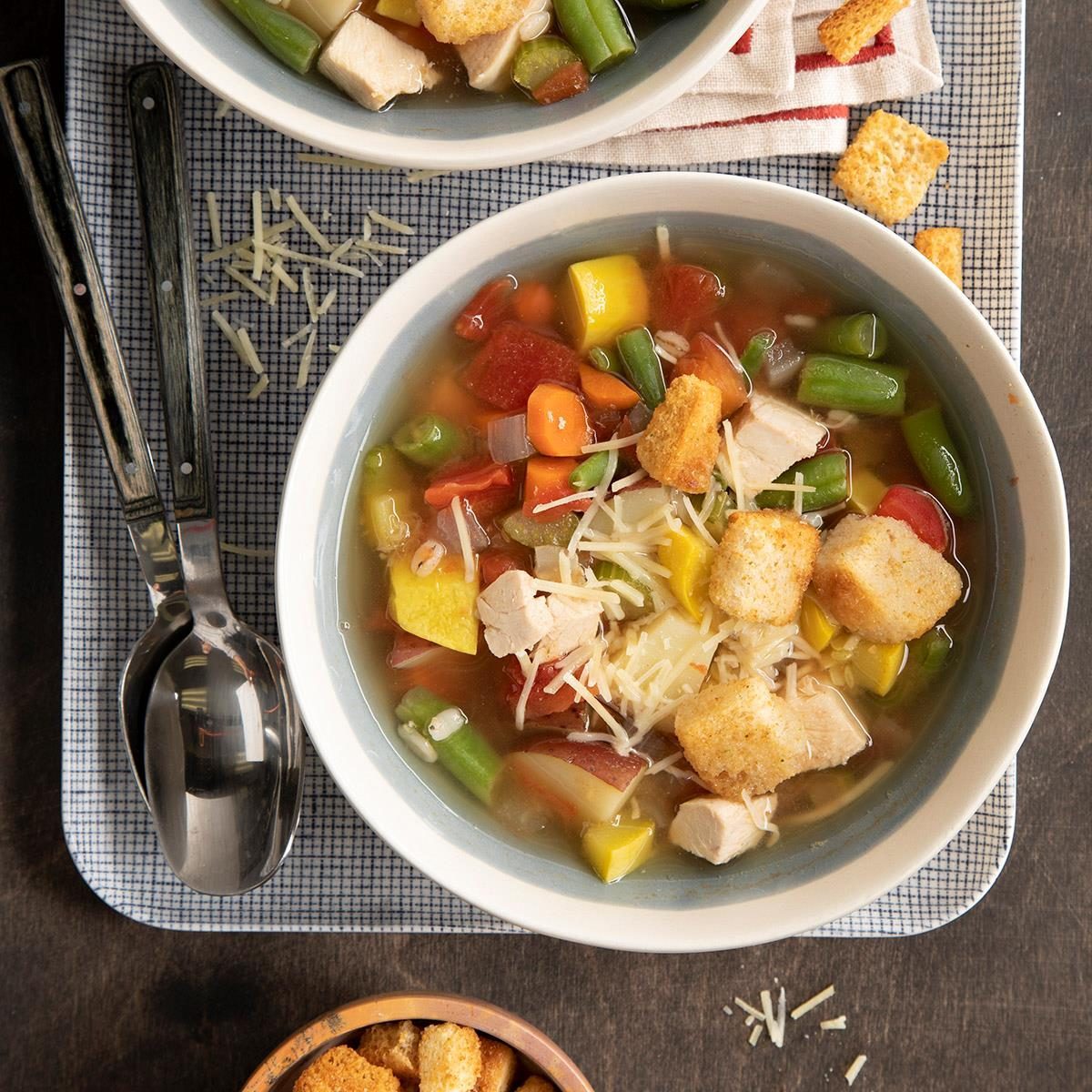 A bowl of vegetable and chicken soup with croutons sits on a checkered tray with two spoons and grated cheese scattered nearby.