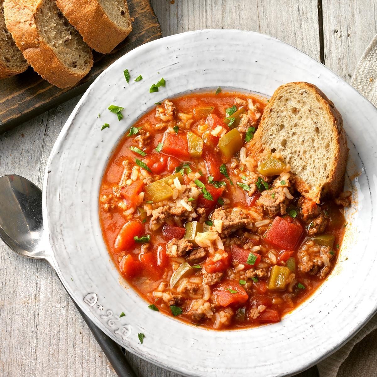 Soup with vegetables and ground meat sits in a white bowl, accompanied by a slice of bread, placed on a wooden table with sliced bread nearby.