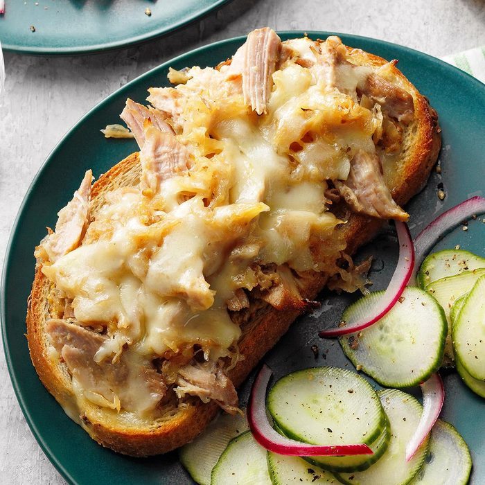 Open-faced sandwich topped with melted cheese and pulled meat, alongside sliced cucumbers and red onions, placed on a green plate.
