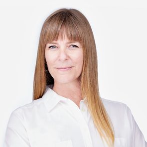 A person with long straight hair and bangs smiles, wearing a white shirt against a plain, light background.