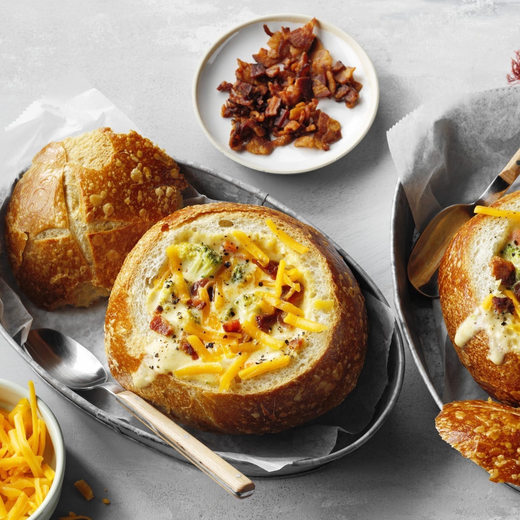 Bread bowls hold creamy soup with bacon and cheese, surrounded by additional bread pieces and a plate of crispy bacon on a gray tabletop.