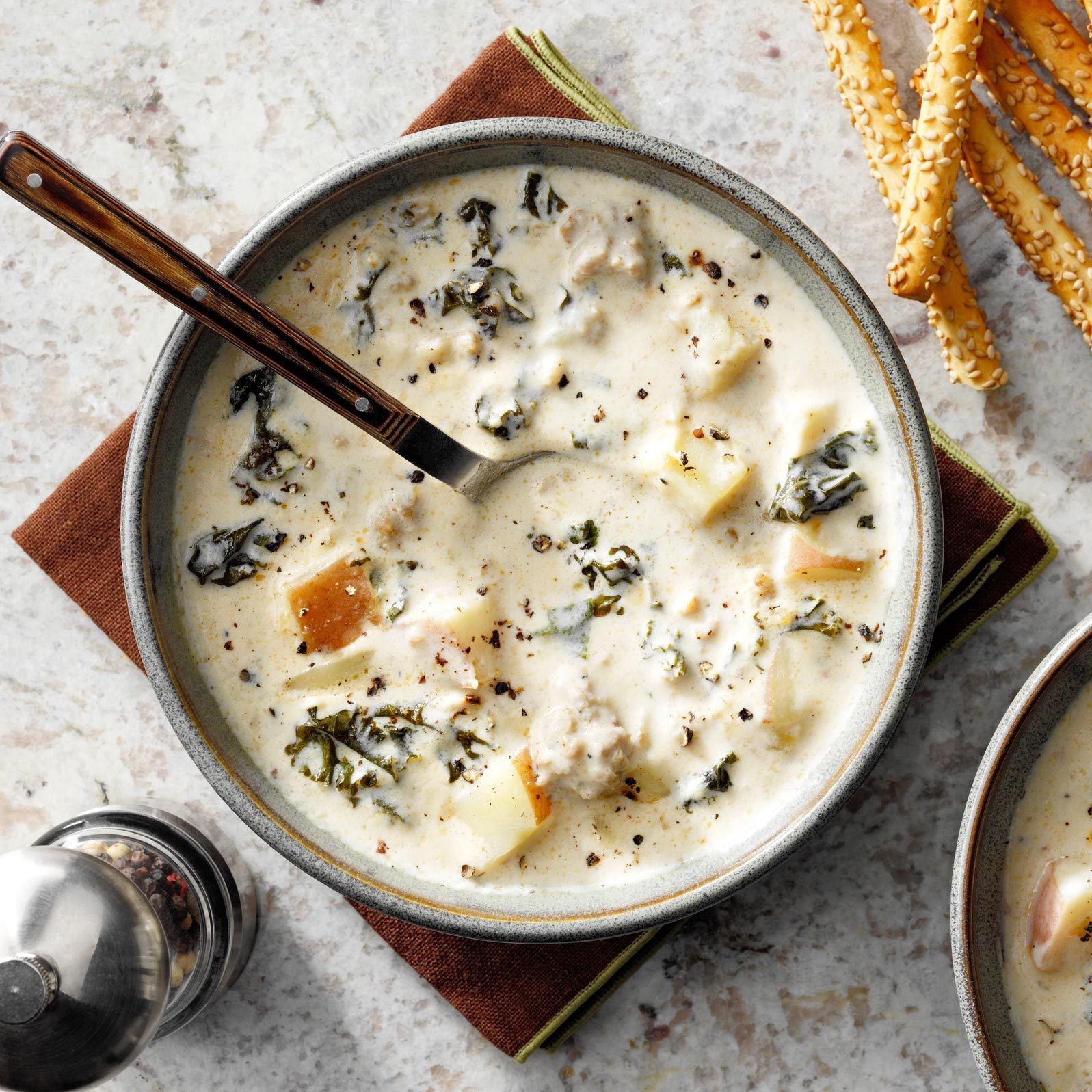 Bowl of creamy soup with kale, potatoes, and ground pepper, on a napkin, beside sesame breadsticks and a pepper grinder on a stone surface.