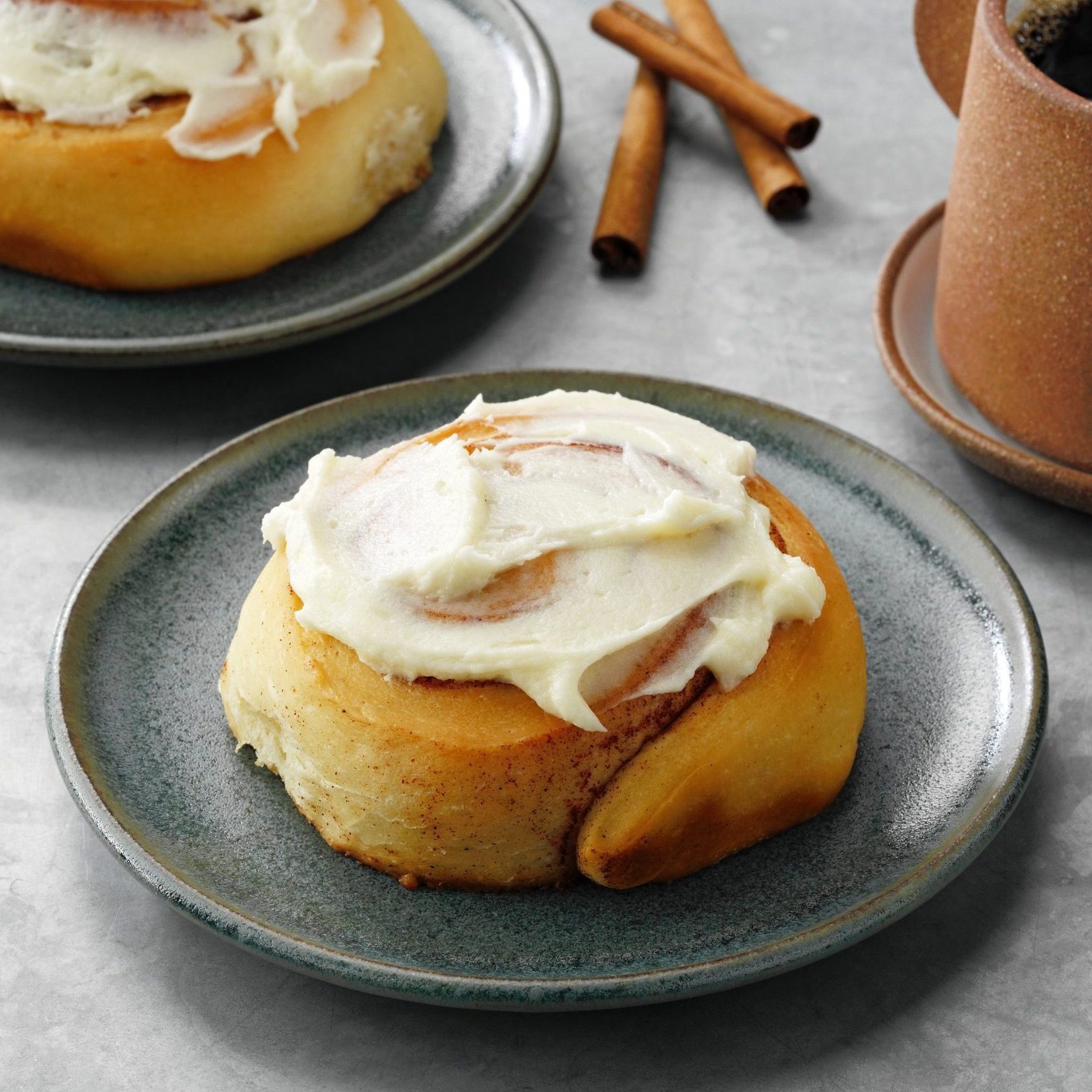 Cinnamon roll sits on a gray plate, topped with white frosting; nearby are cinnamon sticks and a mug on a gray table.