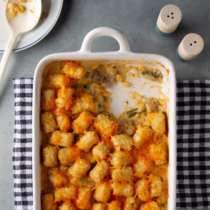 Casserole topped with crispy tater tots in a white dish; a portion is missing on a checkered cloth. Nearby spoon and salt shakers.