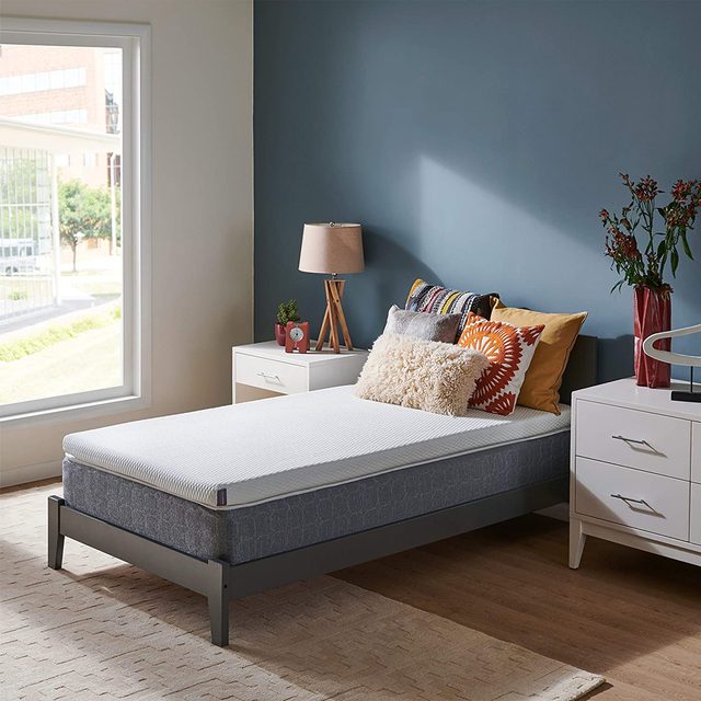Bed with colorful pillows rests in a cozy bedroom, accompanied by wooden nightstands and decorative items, near a large window with sunlight streaming in.