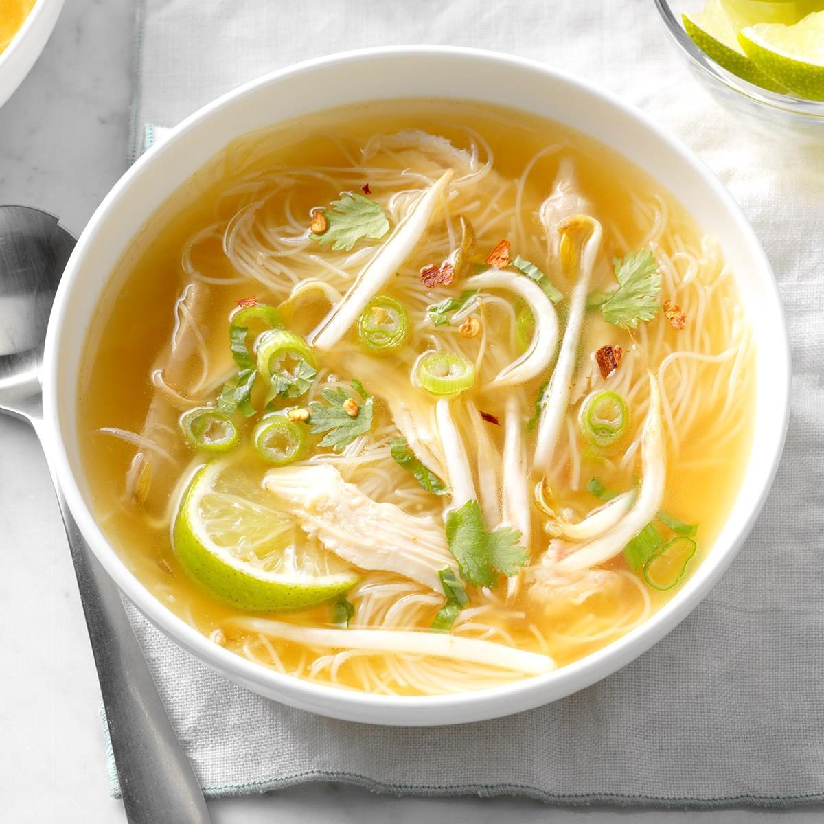 Bowl of noodle soup with chicken, garnished with lime, cilantro, and green onions, placed on a cloth next to a spoon.