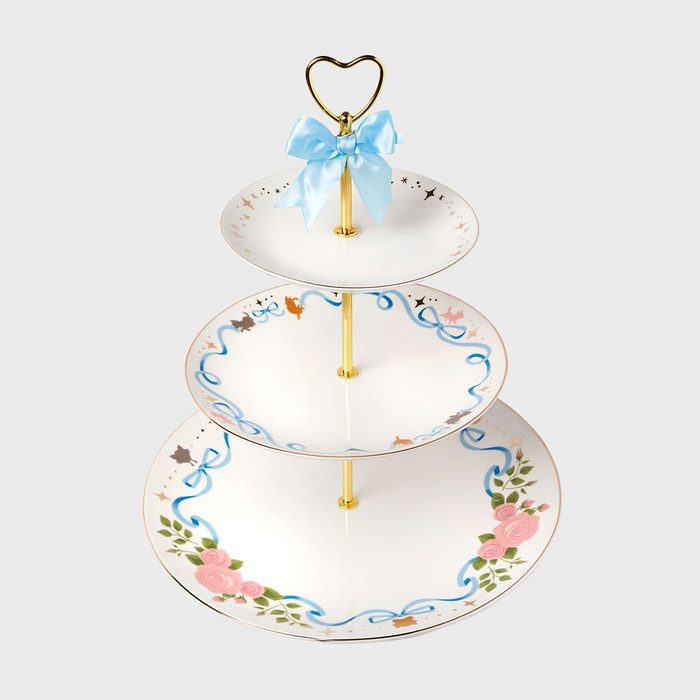 Three-tiered porcelain stand with floral and ribbon design stands adorned by a blue bow on top. Set against a plain background.