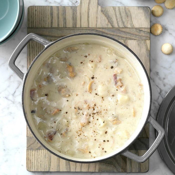 A pot holds creamy soup with chunks, on a wooden board, surrounded by crackers and dishes on a marble countertop.