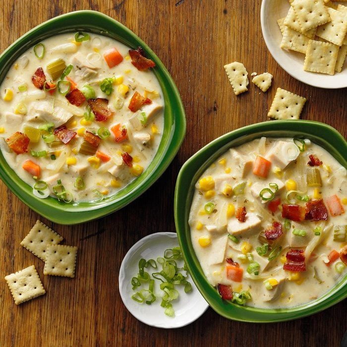 Bowls of creamy soup sit filled with vegetables and bacon, surrounded by scattered crackers on a wooden table.