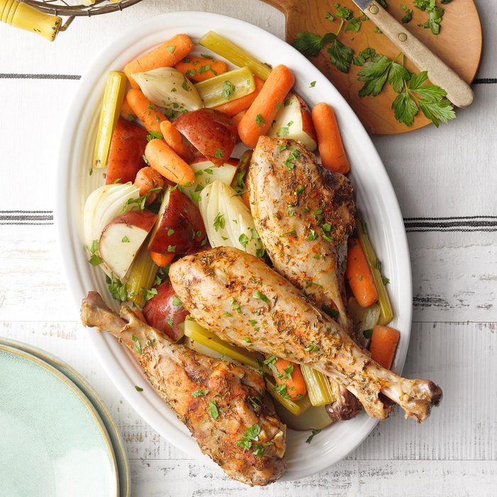 Roasted turkey legs rest on a platter with carrots, potatoes, and onions, garnished with herbs, on a white wooden table.