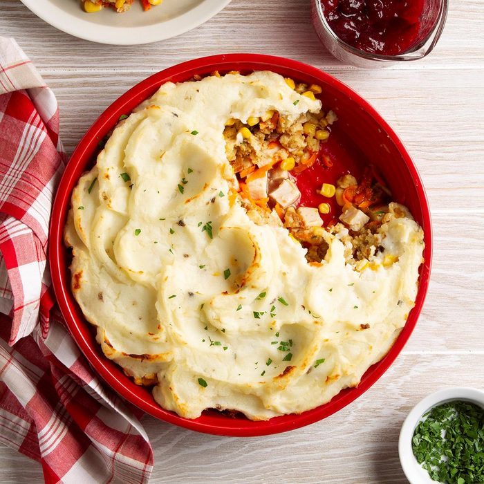 Casserole topped with mashed potatoes, partially eaten, reveals corn, turkey, and stuffing inside, on a table with a red checkered cloth and cranberry sauce.