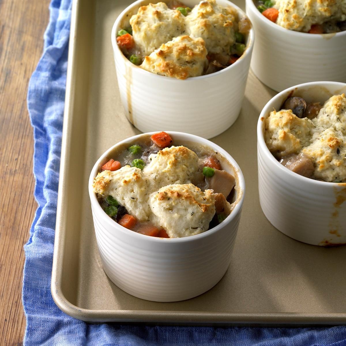 Ramekins hold pot pies topped with biscuit crust, filled with vegetables and sauce, placed on a baking tray with a blue cloth underneath.