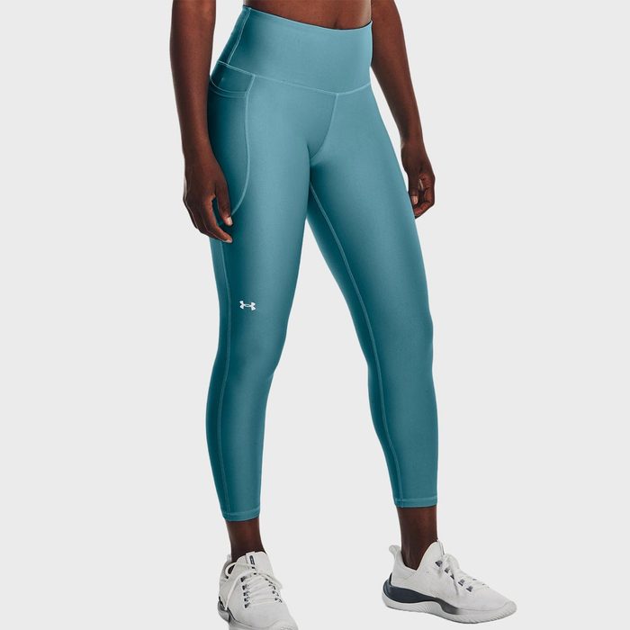 Person wearing teal leggings, standing confidently, with white athletic shoes on a plain backdrop. Under Armour logo visible near the knee.
