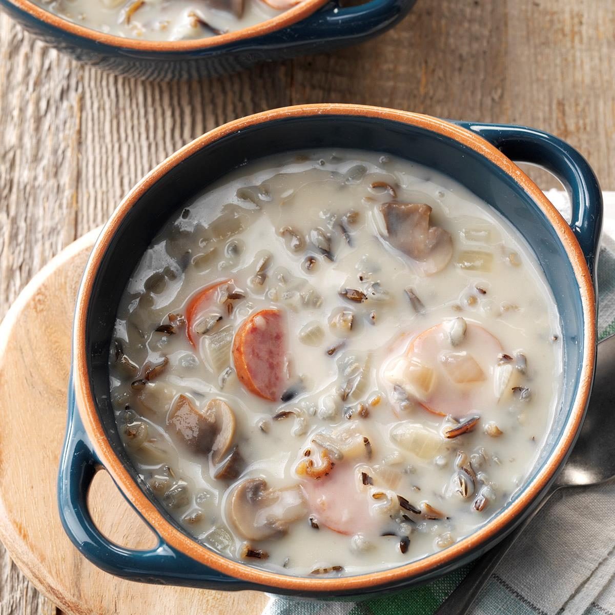 Creamy soup with wild rice, mushrooms, and sliced sausage in a blue bowl; set on a wooden table with a napkin underneath.
