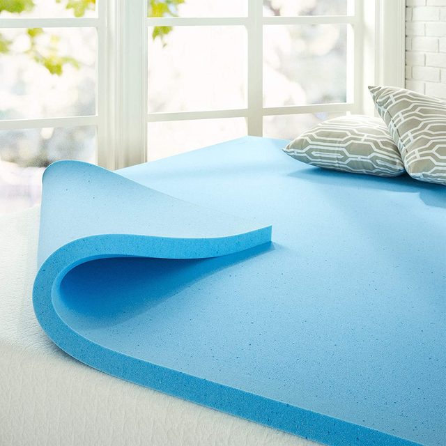 Blue mattress topper lies partially rolled on bed, near patterned pillows; sunlight filters through large windows, creating a bright, airy room.