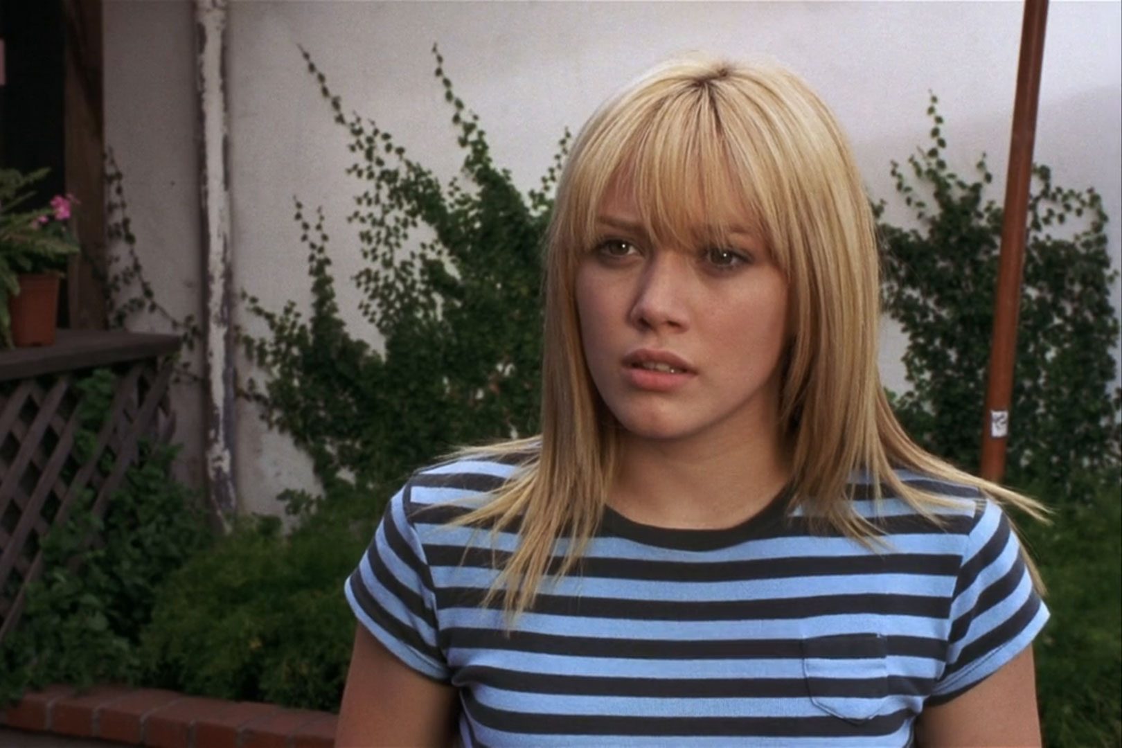 A person with blonde hair looks concerned, standing outdoors with green ivy and a small potted plant in the background.