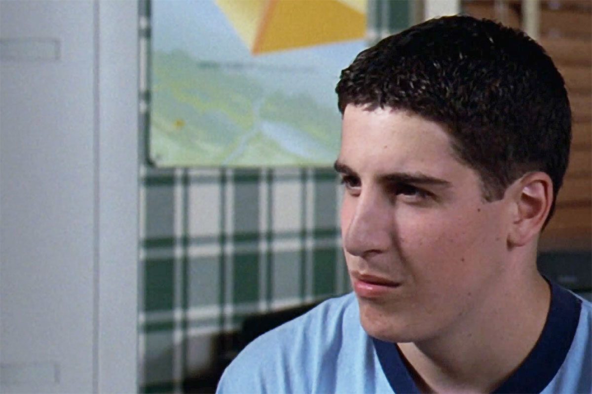 A person looks to the side, wearing a light blue shirt in a room with plaid wallpaper and a colorful poster.