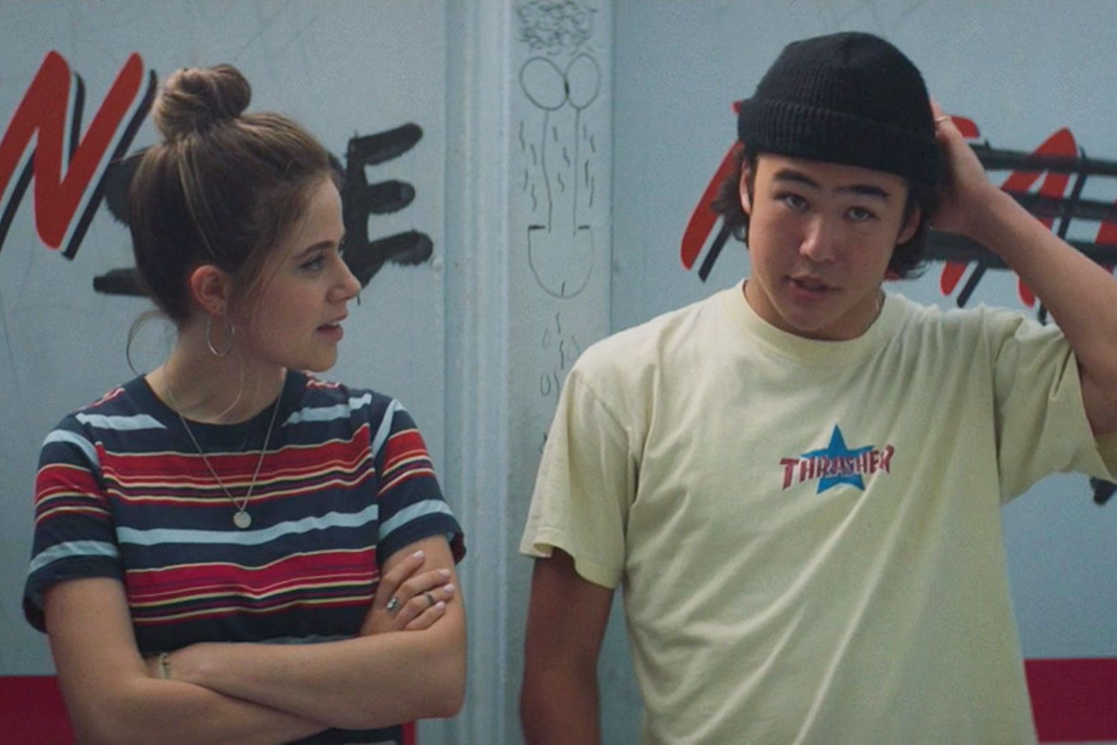 Two people stand against graffiti-marked walls, one talks while the other listens, adjusting a black beanie. Text: "THRASHER" on a t-shirt.