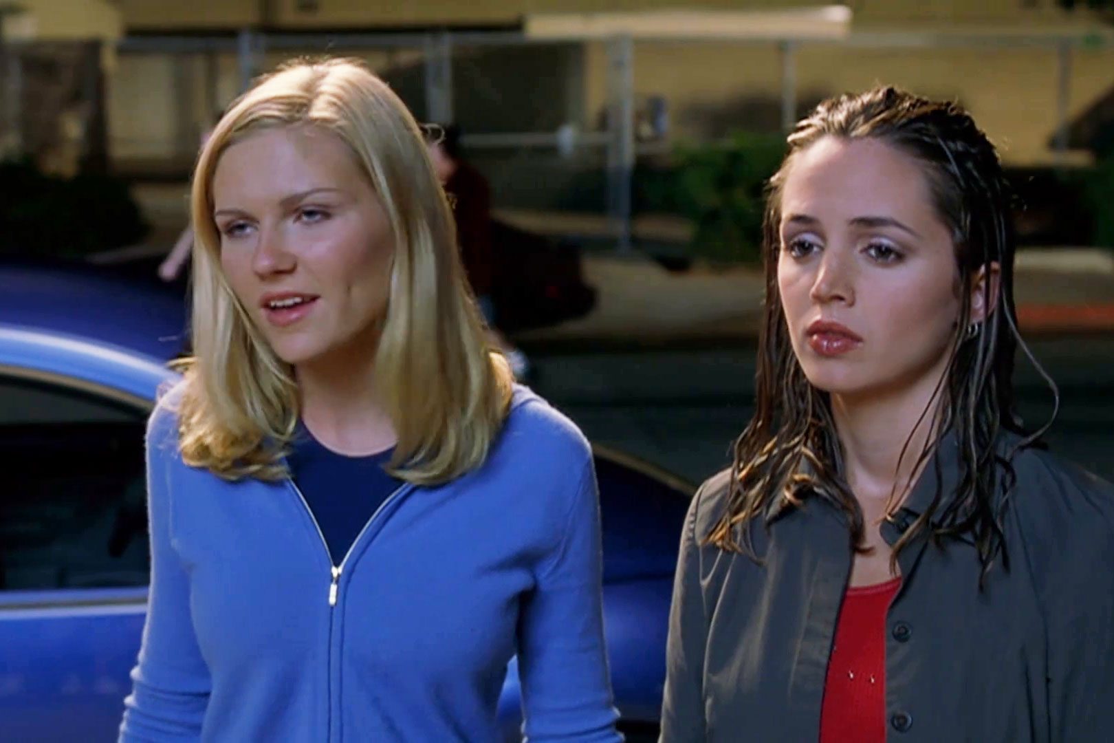 Two women standing, one speaking while the other listens; they are outdoors, possibly near a parked car, with a blurred background.