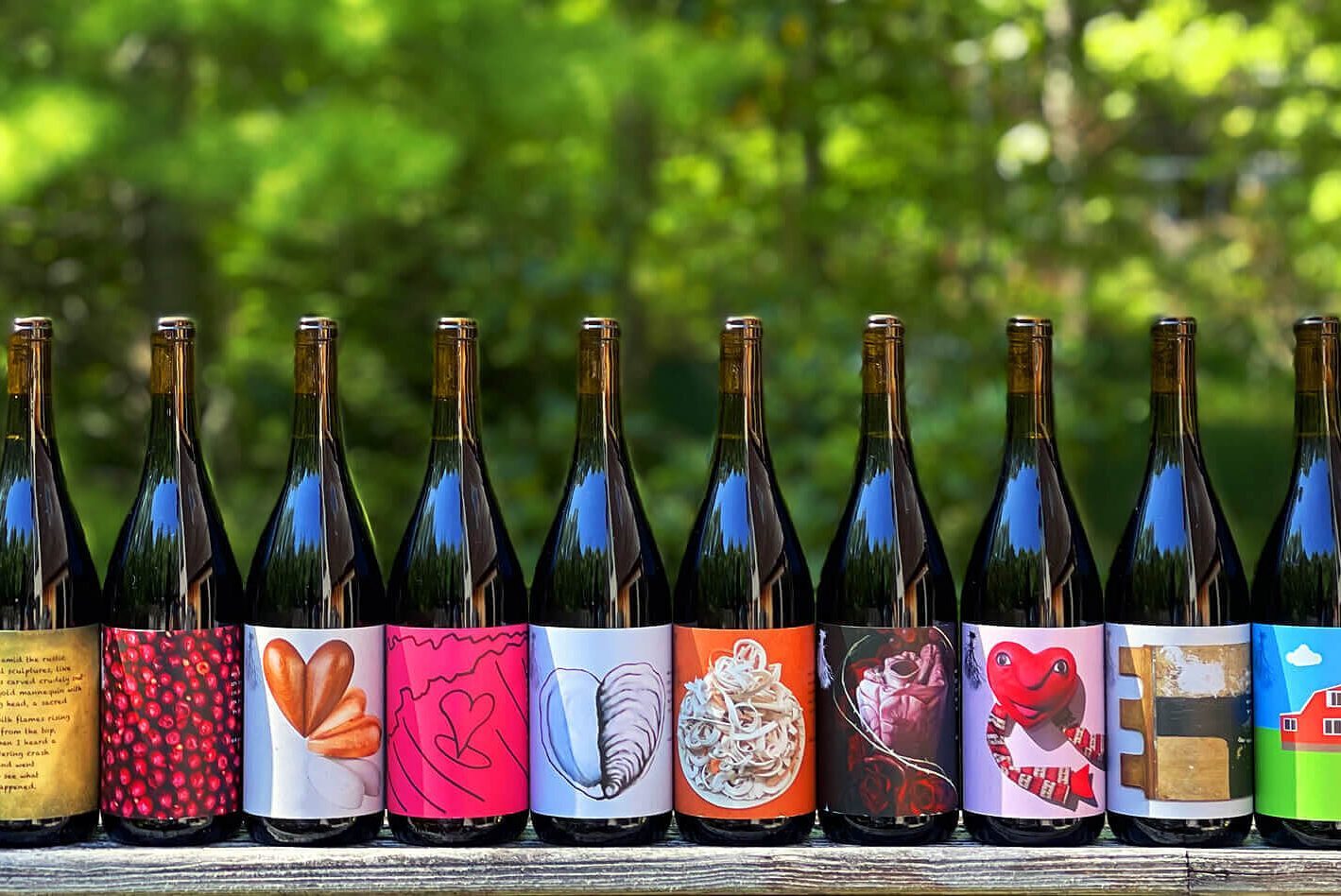 Wine bottles stand in a row, displaying colorful, varied labels, set on a wooden surface against a blurred, green, natural background.