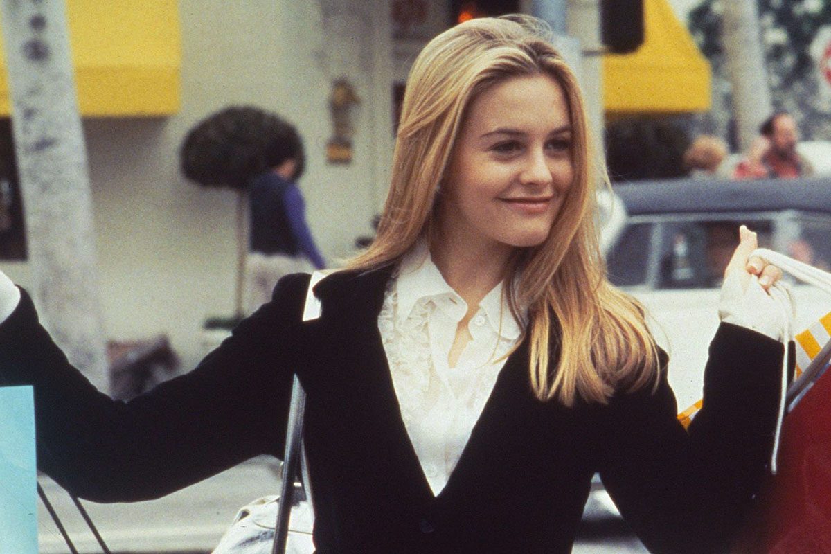 A person smiles while holding shopping bags, standing on a street with blurred background, featuring cars and a yellow awning.