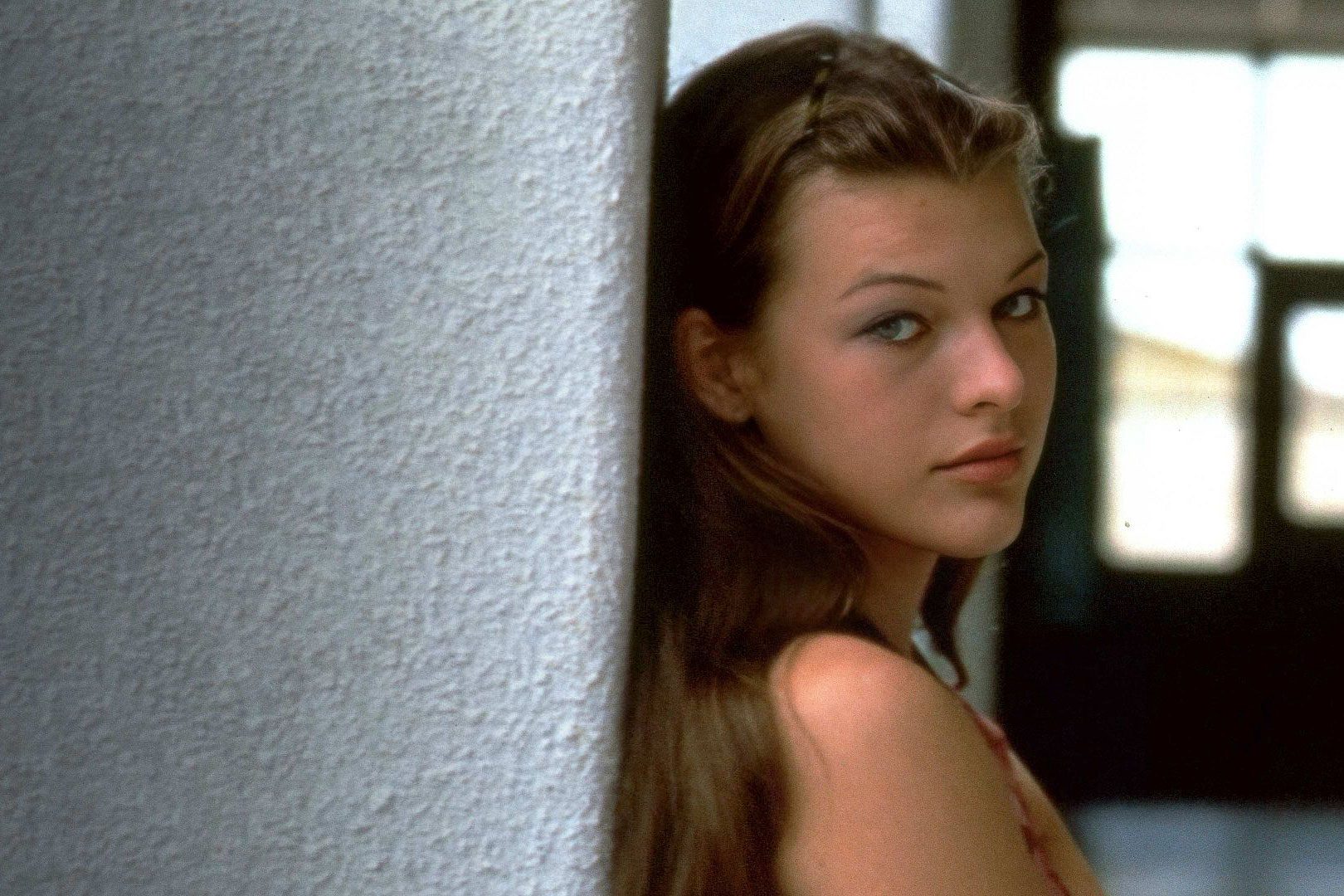 Person leans against a textured wall, looking slightly over their shoulder with long hair, indoors with soft lighting and a blurred background.