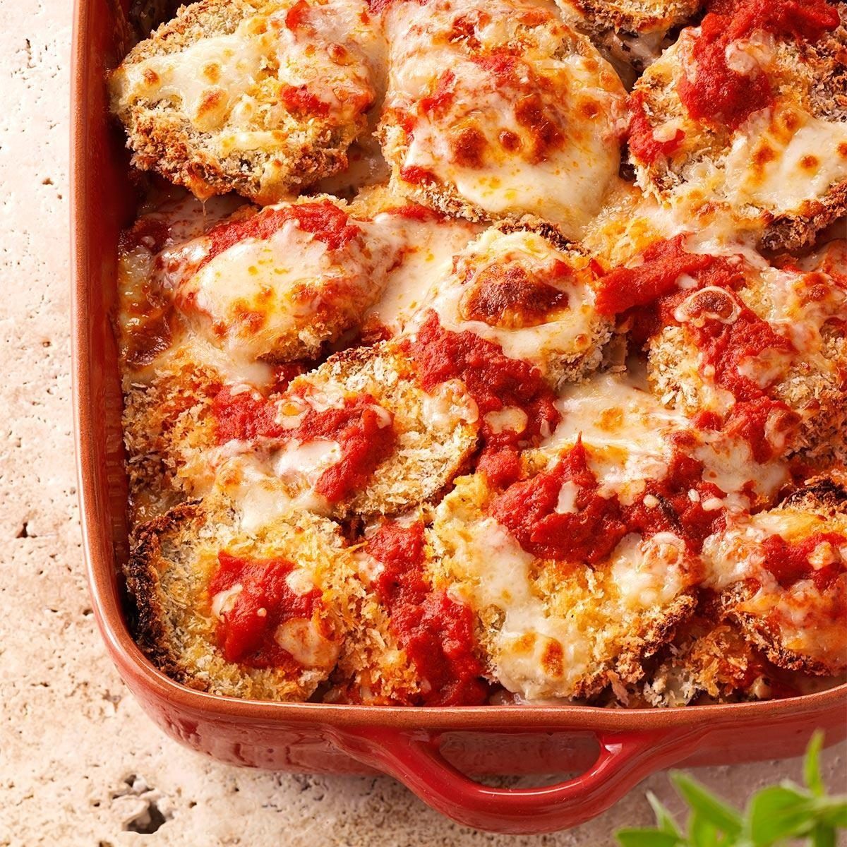 Baked eggplant slices topped with melted cheese and tomato sauce in a red casserole dish on a textured stone surface.