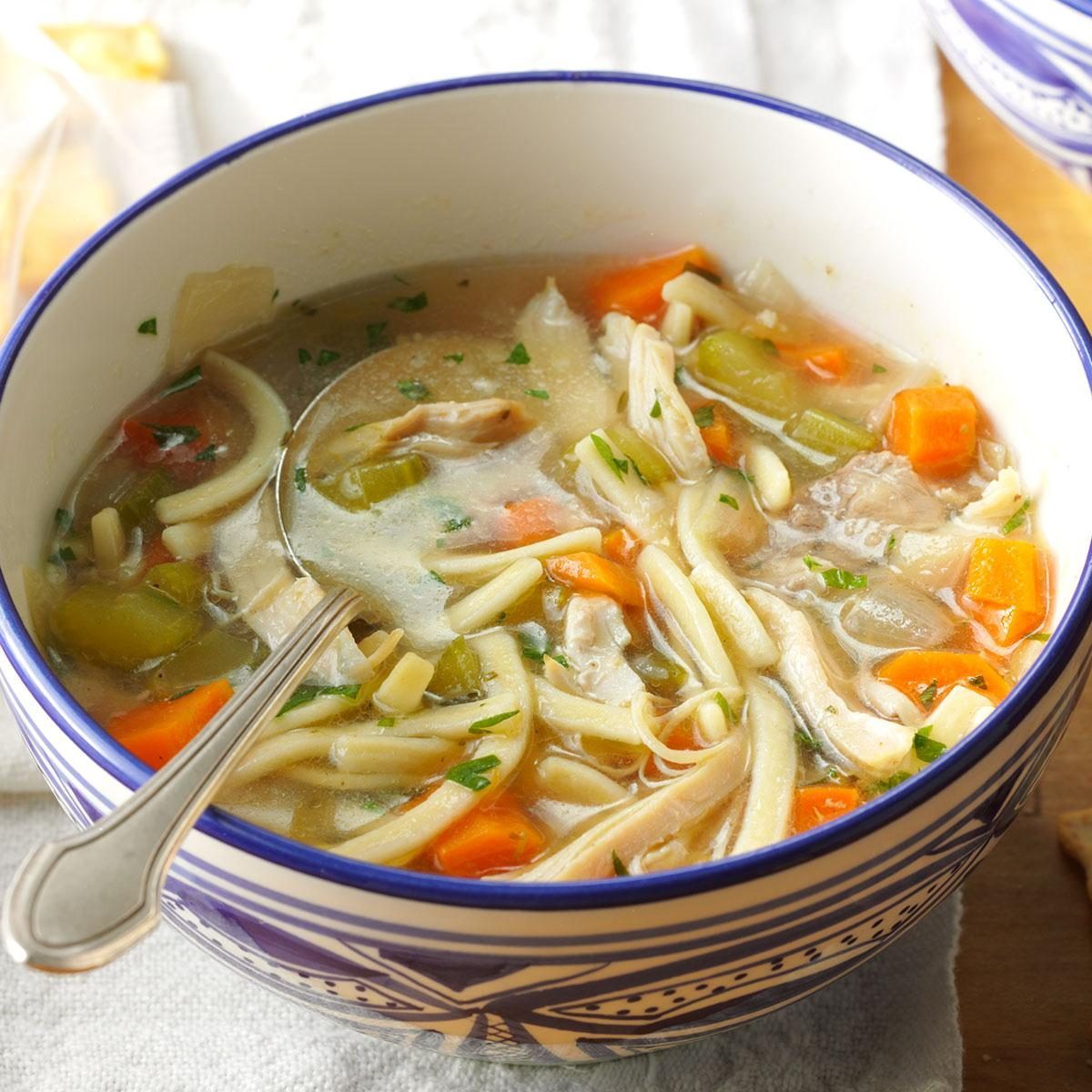 A bowl holds chicken noodle soup with vegetables; a spoon rests inside, surrounded by a patterned rim.