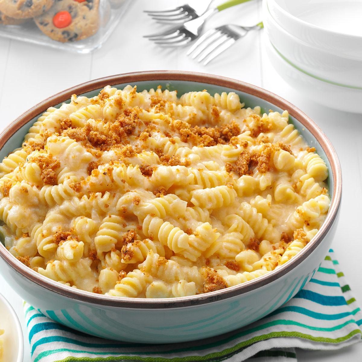A bowl of creamy macaroni and cheese topped with breadcrumbs sits on a striped cloth; forks and stacked bowls are in the background.