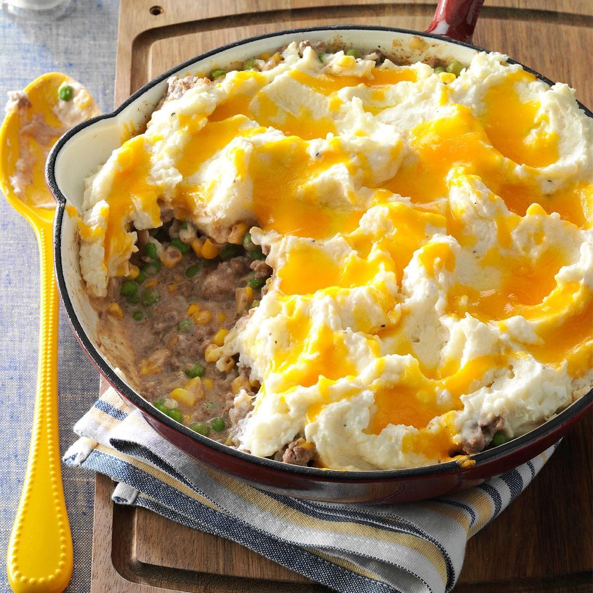 Casserole topped with melted cheese and mashed potatoes, sits on wooden board, with one serving removed, revealing peas and corn inside. Yellow spoon beside it.
