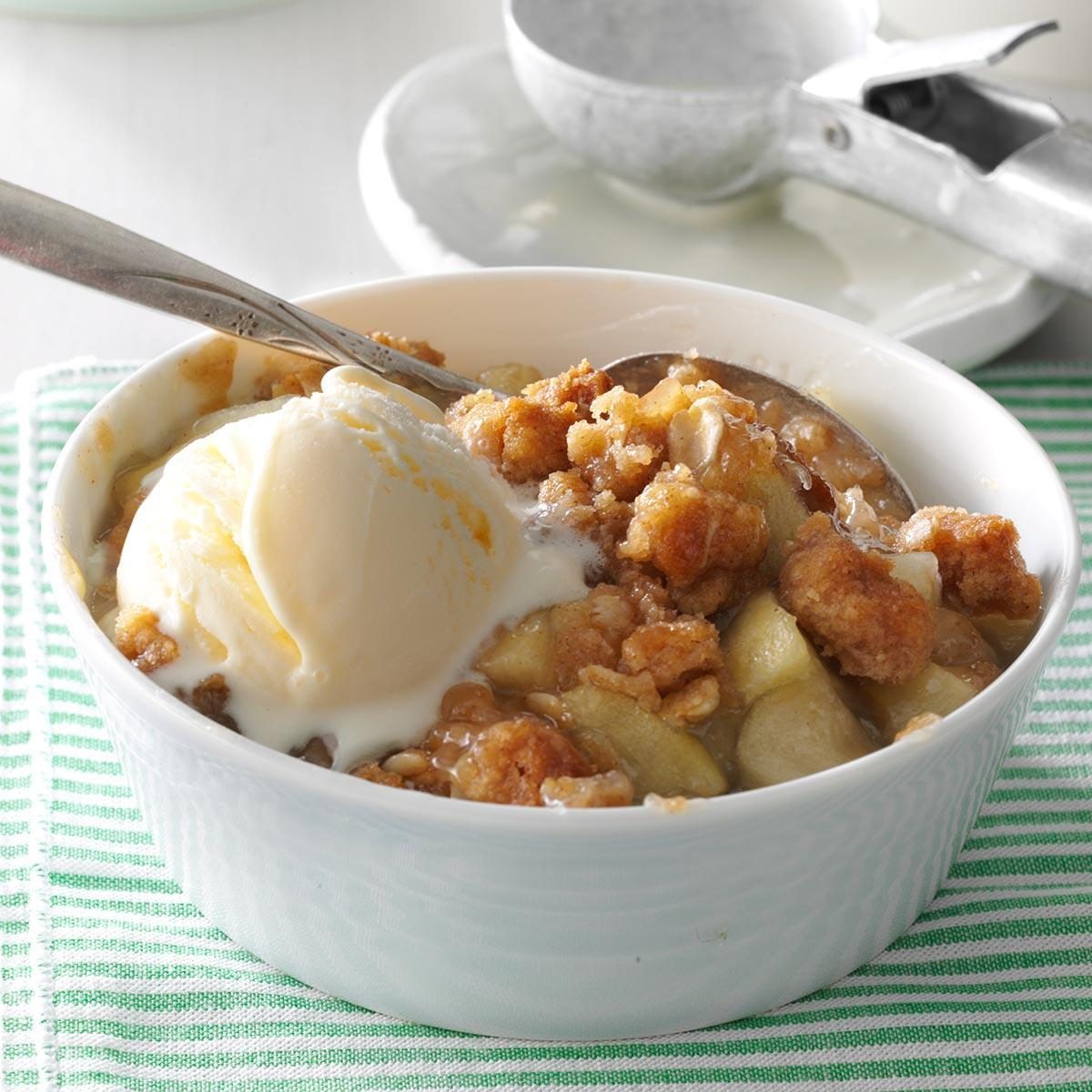 Apple crumble topped with vanilla ice cream sits in a white bowl, resting on a striped cloth. A metal scoop is nearby.