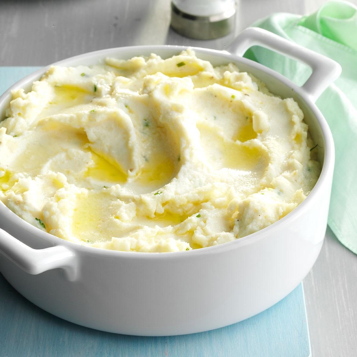 Mashed potatoes are served in a white bowl, drizzled with melted butter, placed on a light blue surface with a green napkin nearby.