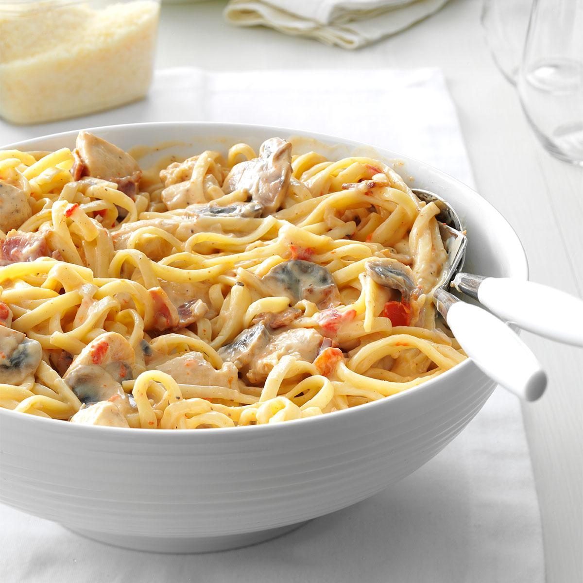 Bowl holding creamy linguine pasta with chicken, mushrooms, and red pepper; set on a white table with a spoon and fork resting inside.
