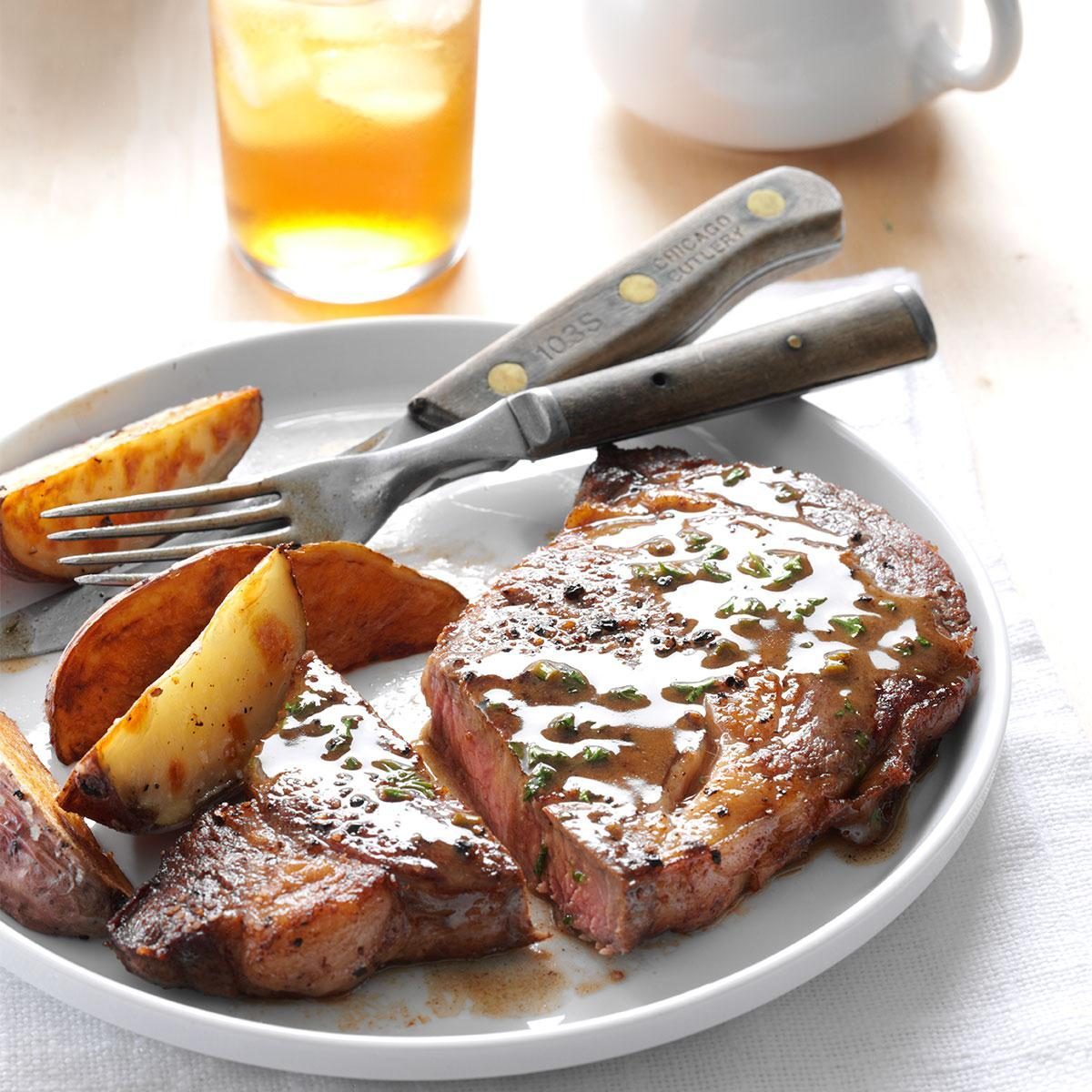 Steak, drizzled with sauce, rests on a plate with potato wedges; utensils are placed atop, an iced tea and teapot are nearby.