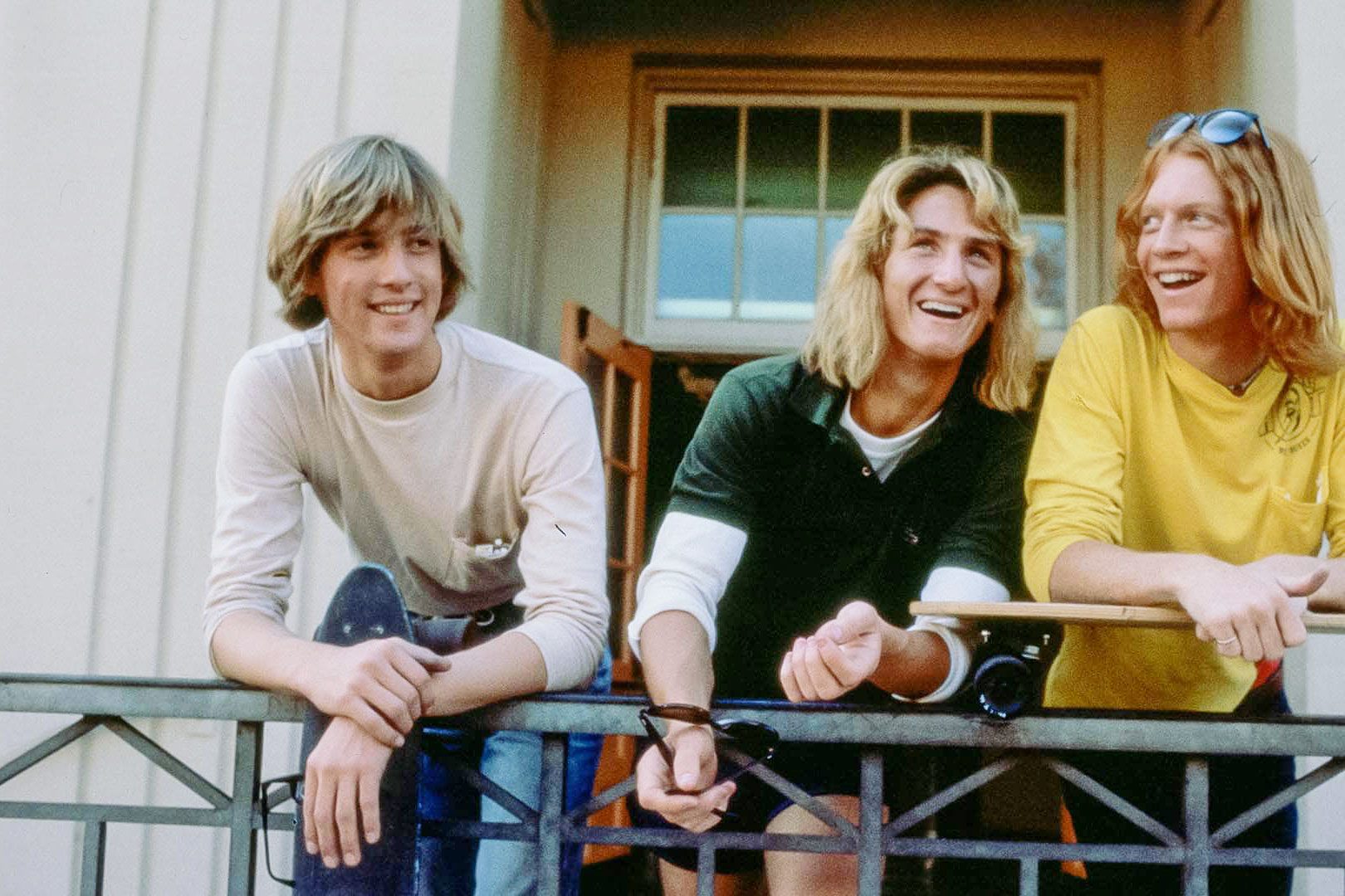 Three young men leaning on a railing smile while holding skateboards, standing outside a building with large windows.