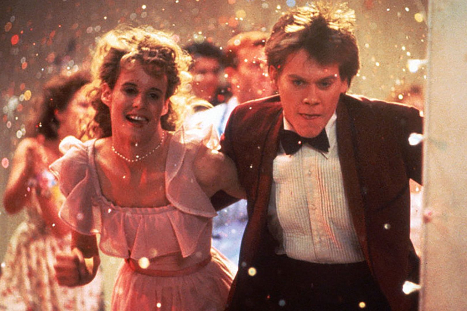 Two people dance energetically at a lively party, surrounded by colorful confetti and a crowd of people in festive attire.