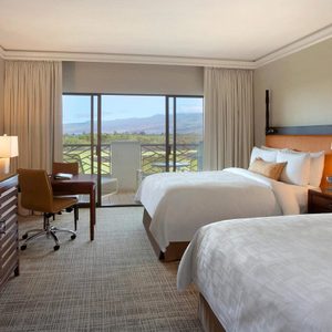 Two neatly made beds sit in a hotel room with a large window revealing a scenic mountain view. A desk and TV are present.