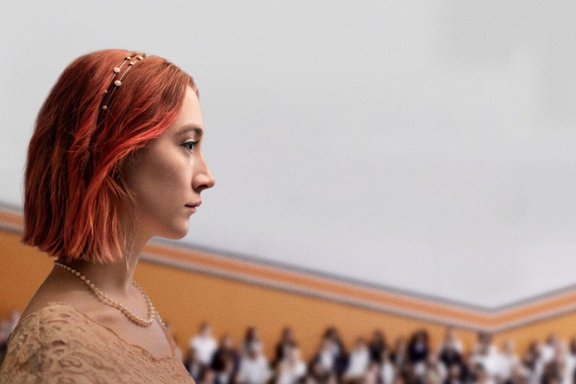 Woman with red hair stands sideways, wearing a lace top and headband, in a room with blurred people seated against beige walls.