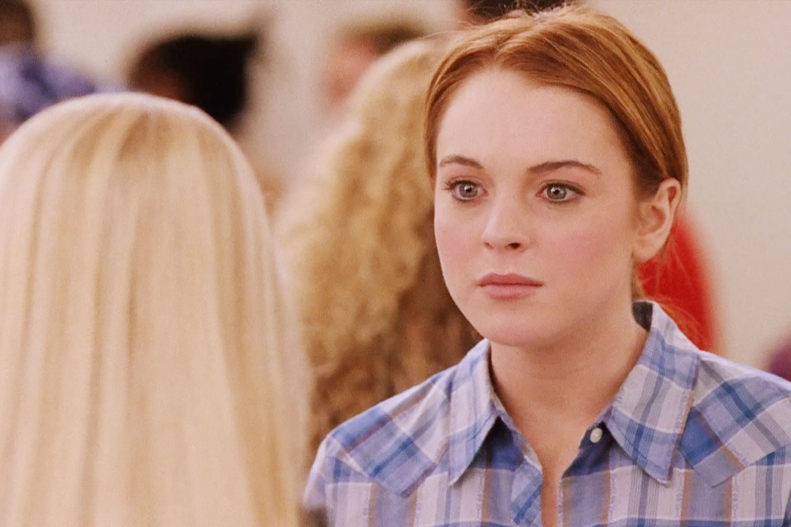 Person stands facing another person in a crowded room, wearing a blue plaid shirt, looking serious.