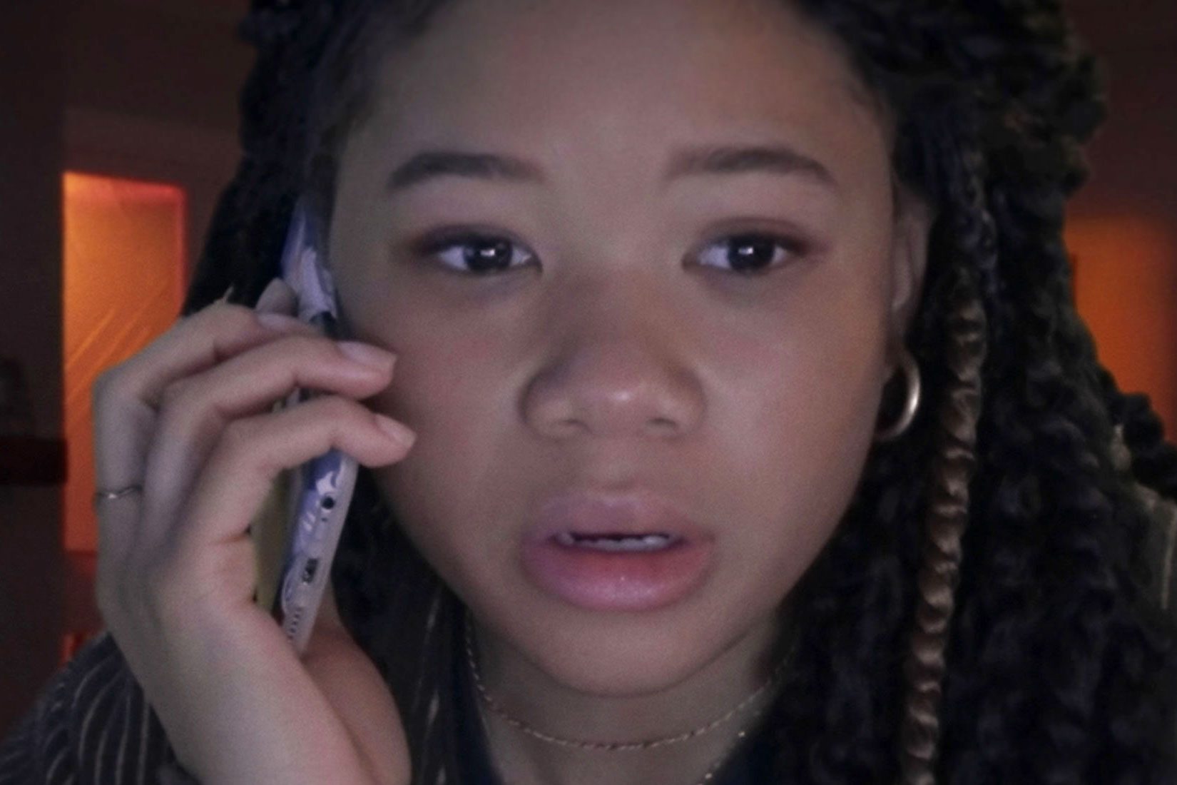 A person holds a phone to their ear, appearing focused in a dimly lit room with soft, warm lighting in the background.