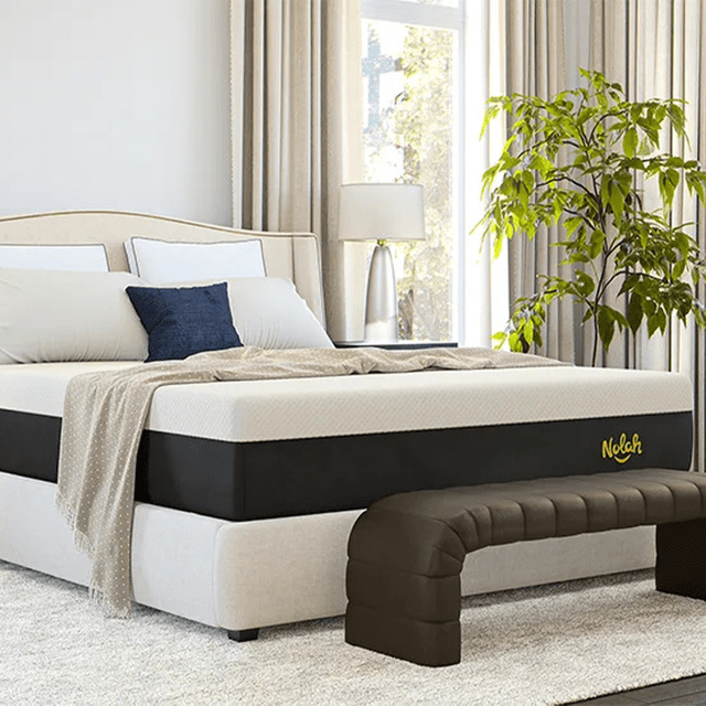 Bed is neatly made with pillows and a blanket, situated in a bright, spacious bedroom with a plant and beige curtains nearby.