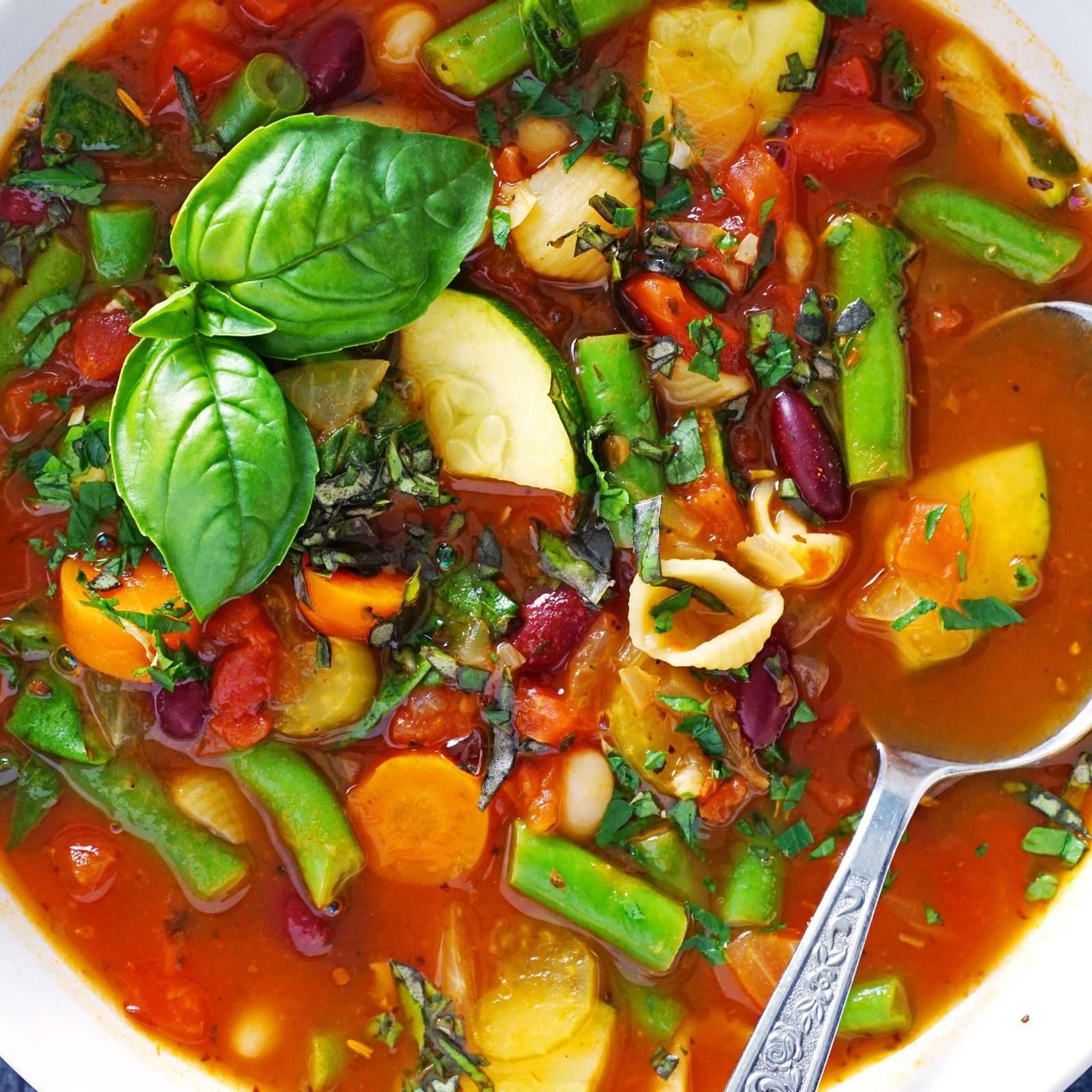 Soup brimming with vegetables and beans, garnished with basil, in a bowl. A spoon rests in the vibrant broth.