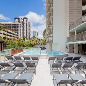Lounge chairs sit empty beside a rooftop pool, surrounded by palm trees and high-rise buildings under a clear blue sky.