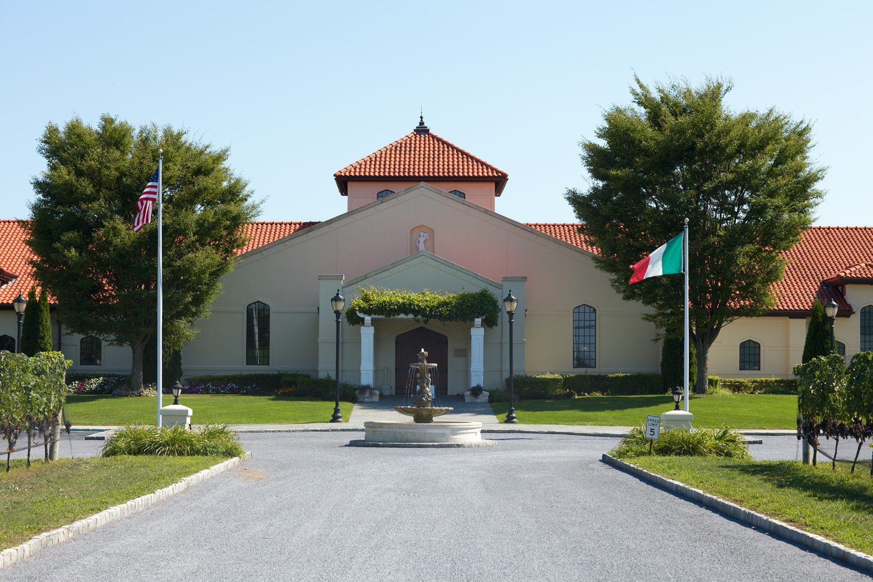 Fountain spouts water in front of a beige building with red roof, surrounded by trees and flags on poles.