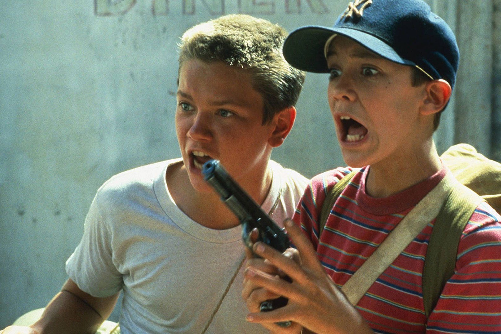 Two boys, one holding a gun, appear frightened, standing in an outdoor setting with a concrete wall in the background.