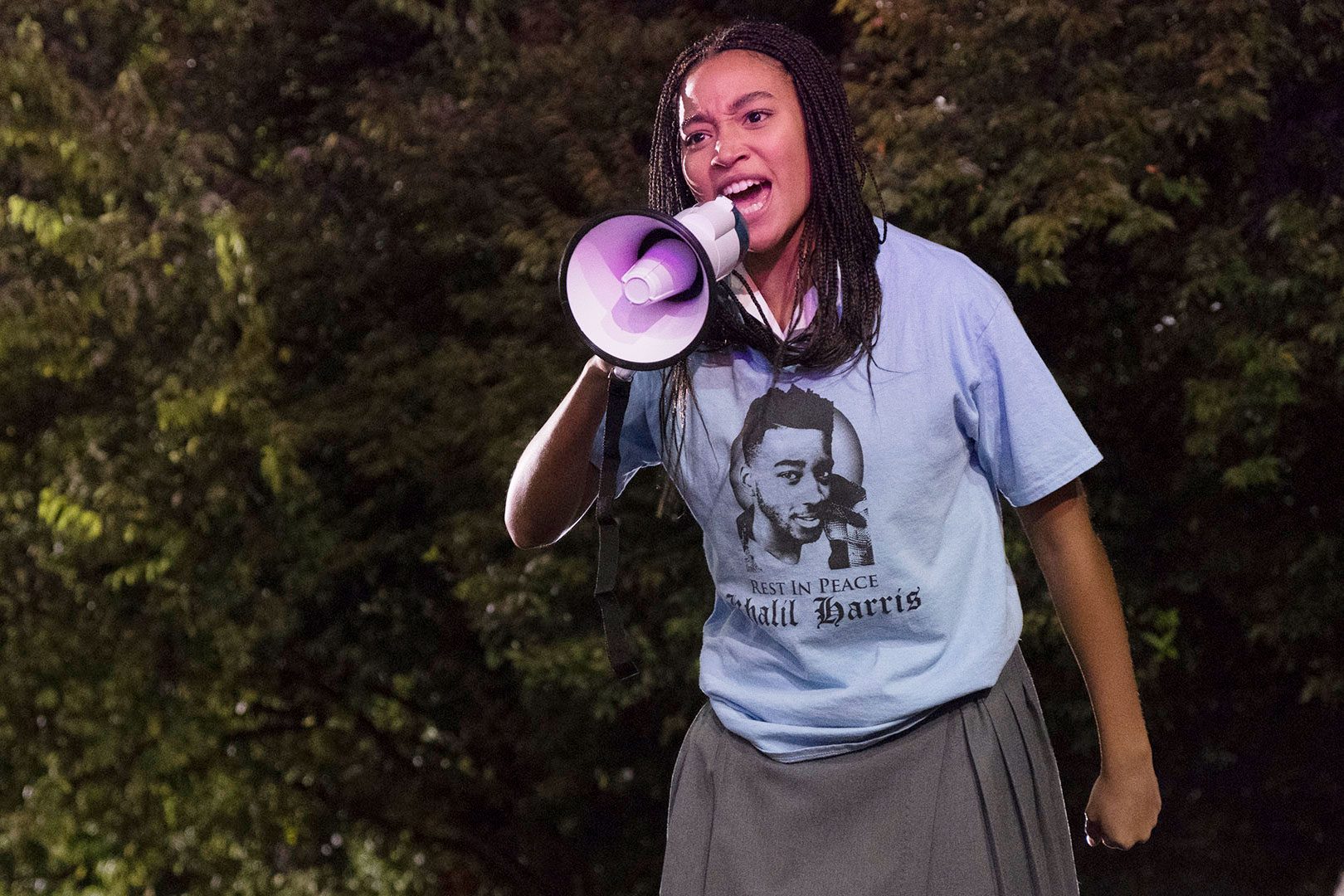 Person holding megaphone, speaking passionately in a park. T-shirt reads, "Rest in Peace Khalil Harris," with a portrait. Dense foliage in the background.