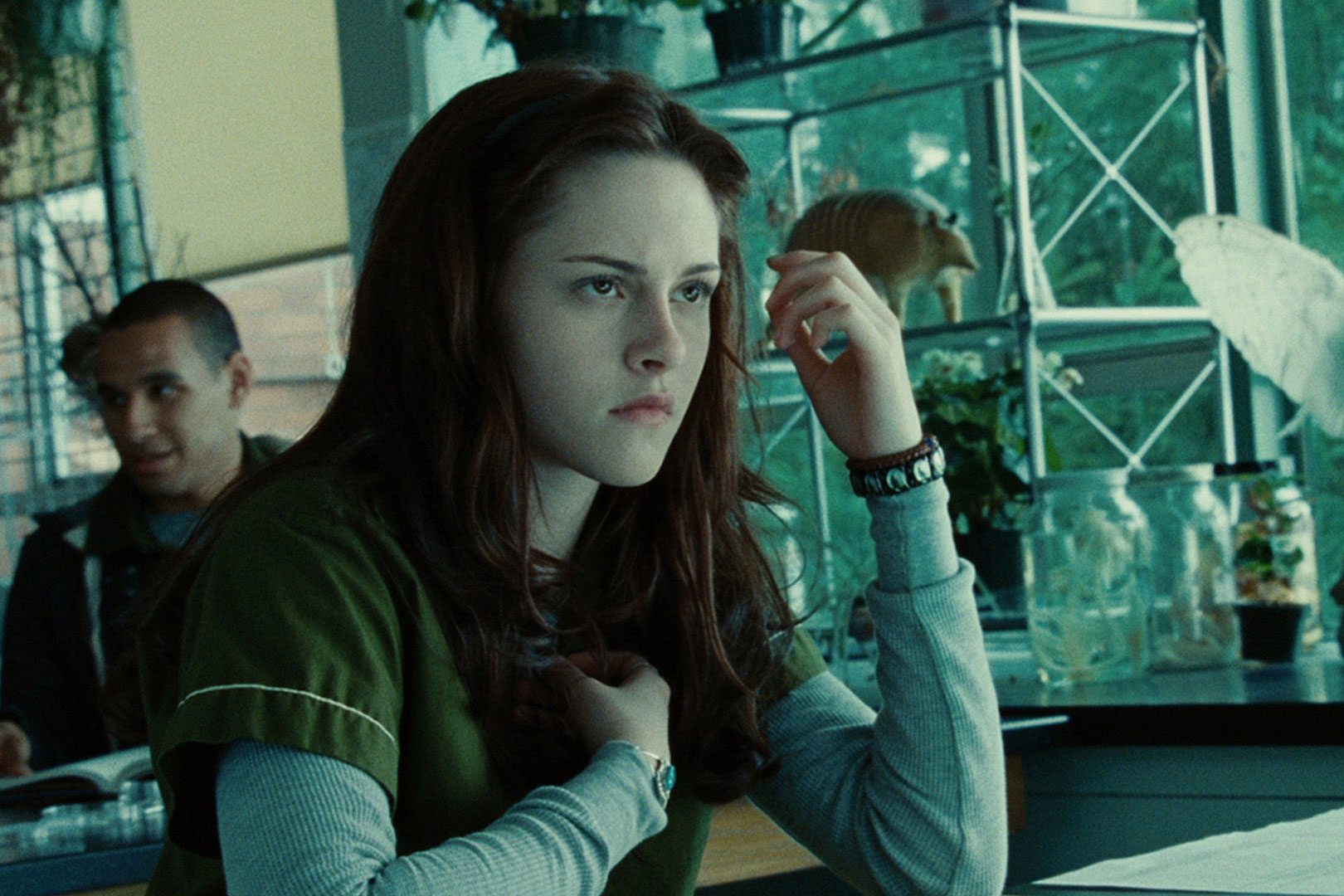 A person sits thoughtfully at a desk surrounded by jars, plants, and shelves in a green-lit room.