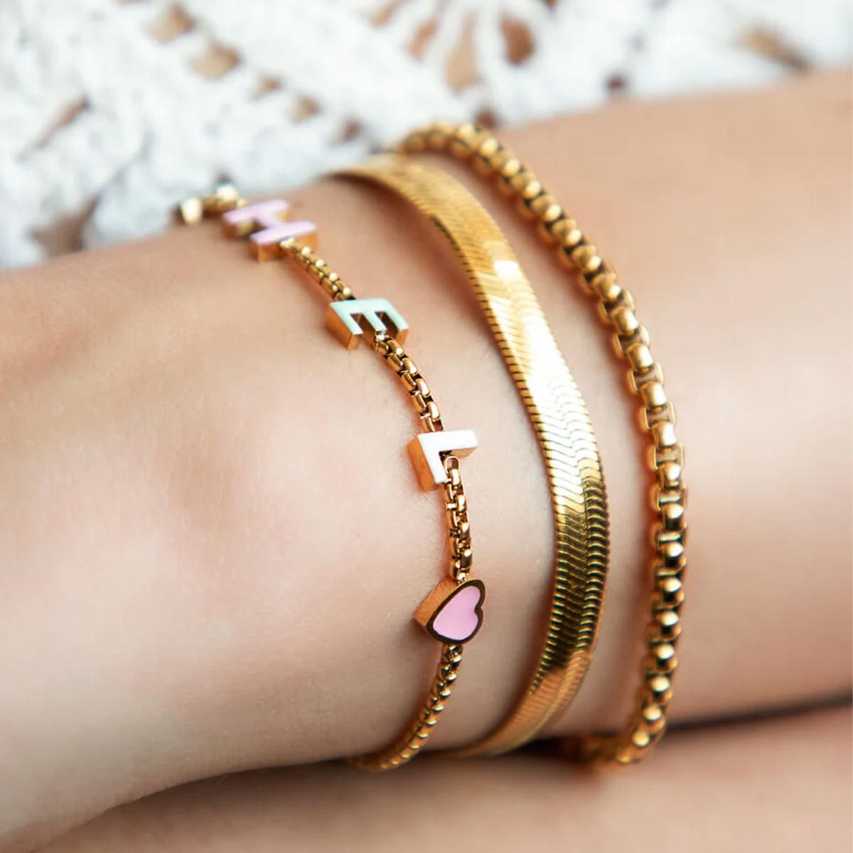 Gold bracelets adorn wrist; one features letters and a pink heart. Background has lace fabric texture.