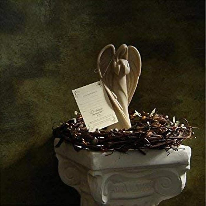 Wooden angel rests in a twig nest atop a pedestal, accompanied by a card against a dark, textured background.